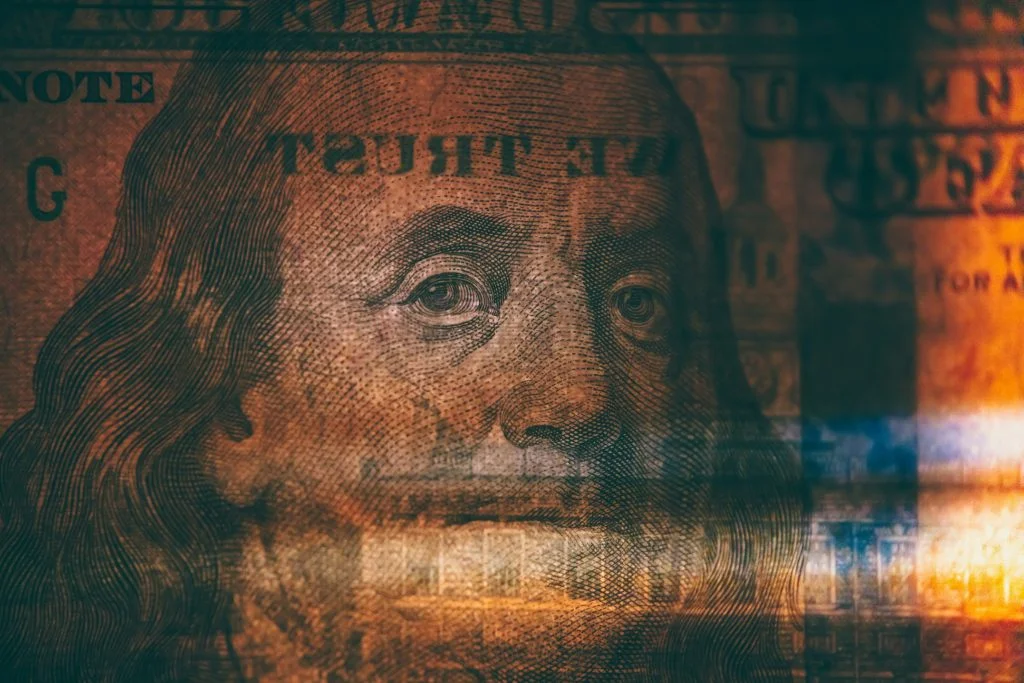A close-up of Benjamin Franklin's portrait on a $100 bill, with a dramatic warm orange and amber color grade and a light leak effect across the right side of the frame.