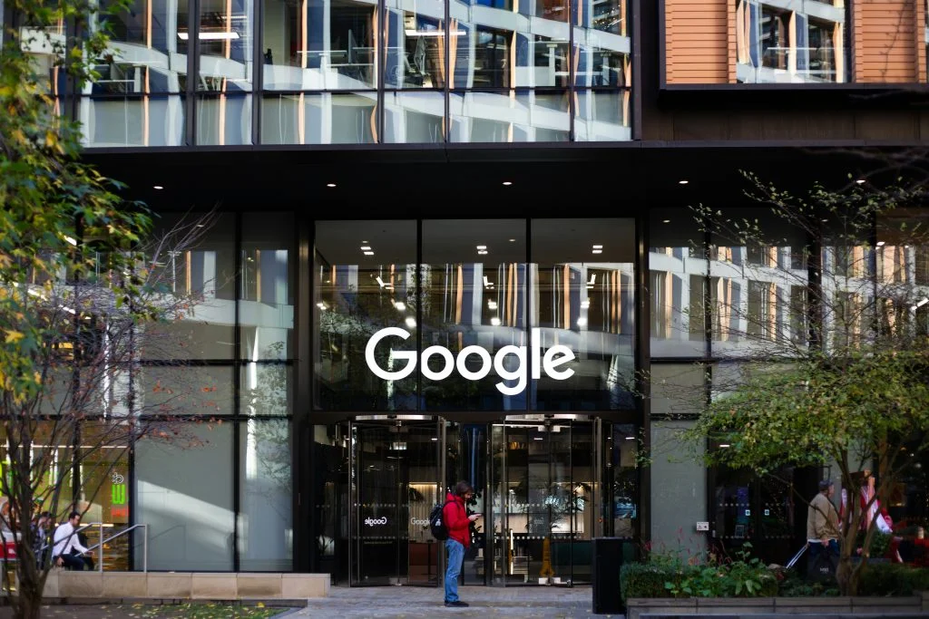 Google logo above the glass entrance to a modern office building, with pedestrians and trees outside.
