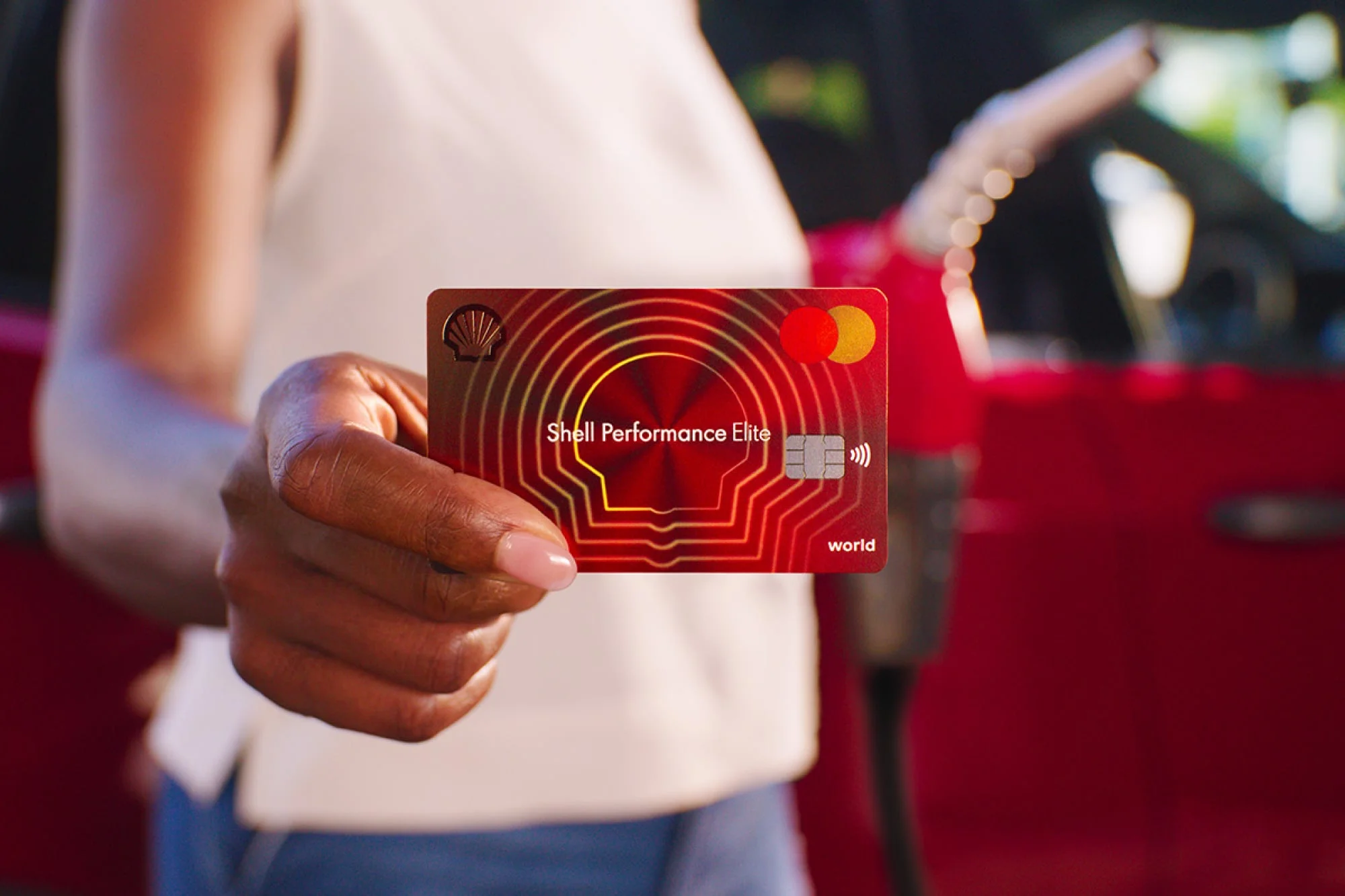 Woman holding up a Shell Performance Elite credit card while holding a gas pump in the background