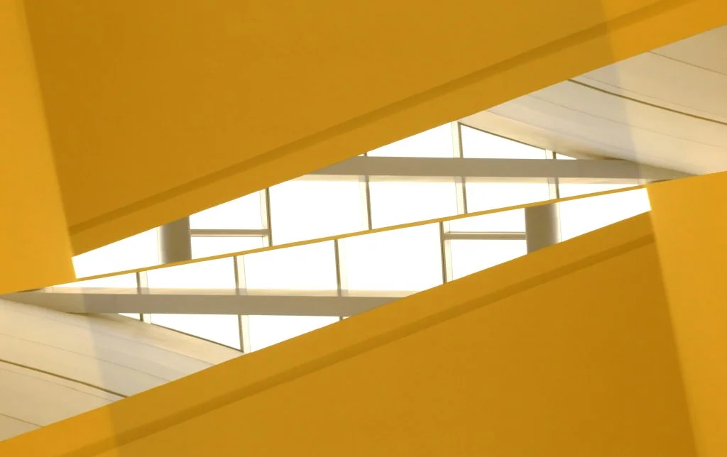 Abstract view up through bold yellow angled beams to a white skylight grid and curved ceiling panels.