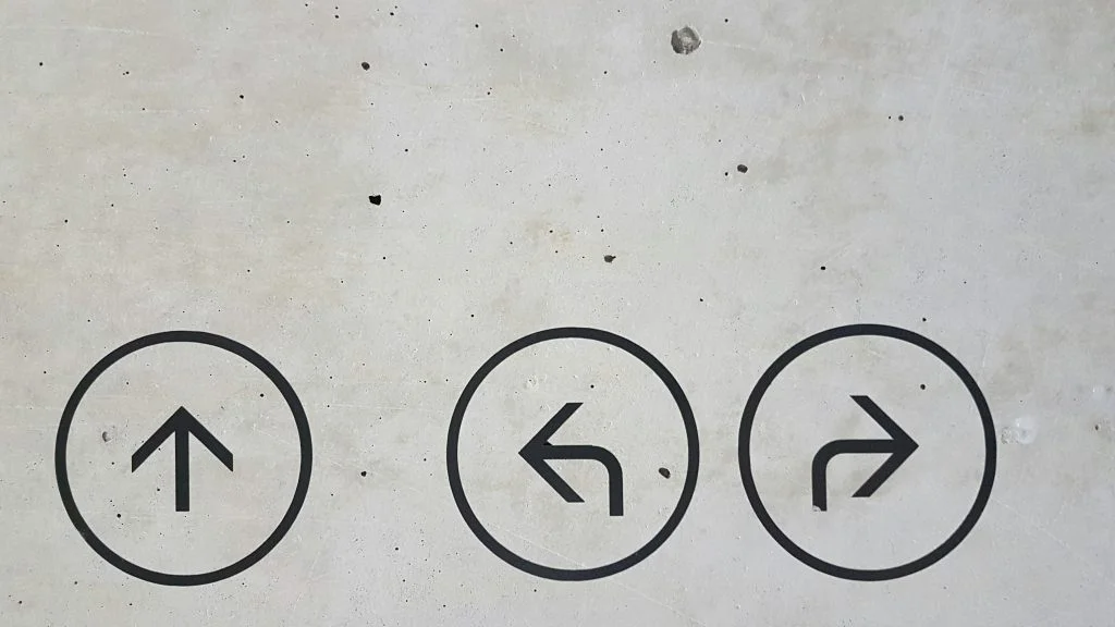 Three black circular directional signs on a gray concrete wall, showing arrows pointing straight ahead, turning left and turning right.